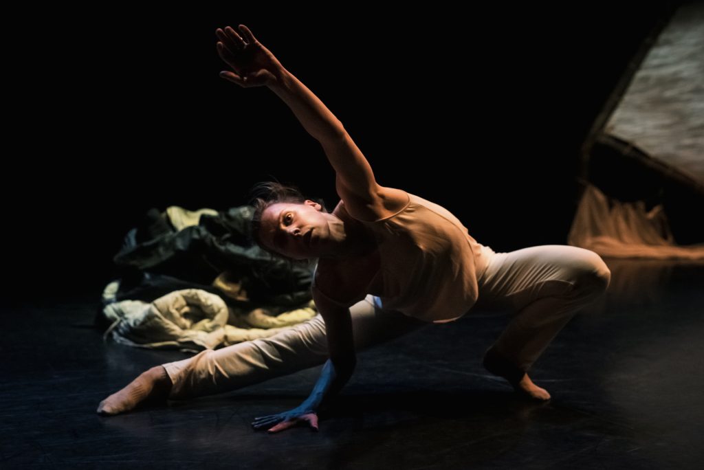 Dance Alexis Fletcher in a low stretched position on stage with dark lighting. 